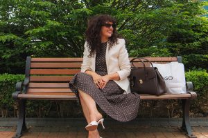 Kate Lewis, Merry Hill's Head of Seasons for spring summer fashion, wears a polka dot outfit and blazer, sitting on a bench with foliage in the background